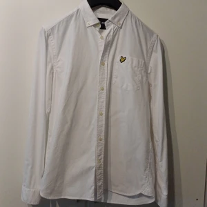 Vit skjorta från Lyle & Scott - Klassisk vit skjorta från Lyle & Scott med lång ärm och bröstficka. Skjortan har knappar framtill och en broderad gul logga på bröstet. Perfekt till många olika tillfällen och enkel att matcha med andra plagg.