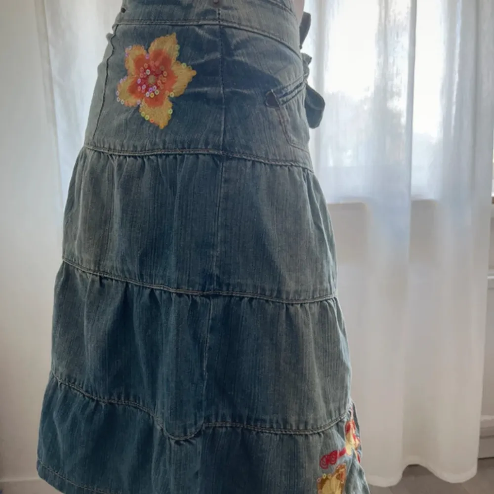 Säljer en blå jeanskjol med volanger och broderade blommor i gult och rött. Kjolen har knytband i midjan och en liten ficka framtill. Perfekt för dig som gillar en avslappnad och söt stil.. Hameet.