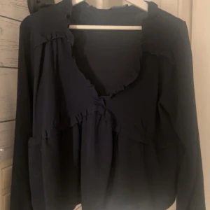 Mörkblå blus med volangdetaljer - Säljer en mörkblå blus med långa ärmar och volangdetaljer längs halsringningen och över bröstet. Blusen har en lös passform och en feminin v-ringning. Perfekt att styla till jeans eller kjol.