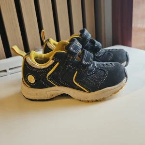 Svarta och gula sneakers med kardborre - Säljer ett par svarta sneakers från Timberland med gula detaljer och vit sula. Skorna har två kardborreband för enkel påtagning och en sportig look. Perfekta för aktiva dagar och lek. Fraktkostnaden beror på adressen.