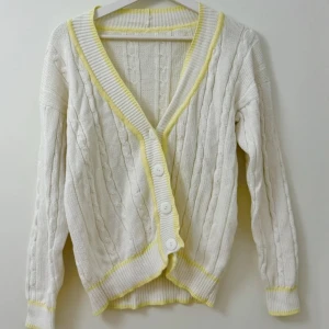 Vit kofta med gula detaljer - Säljer en vit kabelstickad kofta med ljusgula ribbade kanter och stora vita knappar. Koftan är tillverkad i 100% bomull och har lång ärm samt v-ringad hals. 