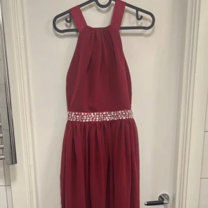 Vinröd långklänning med strassdetaljer - Säljer en vinröd långklänning med breda axelband som korsas i ryggen och markerad midja med strassdetaljer. Klänningen har ett elegant fall och är perfekt för bal eller fest. Materialet känns mjukt och har ett fint skimmer. OBS har en missfärgning på en av banden där bak!!