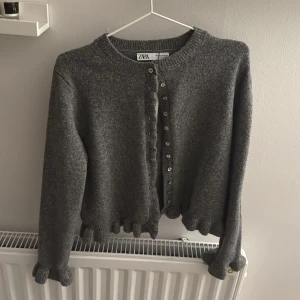 Grå stickad kofta från Zara - Mysig grå stickad kofta från Zara med volangkant nertill och vid ärmslut. Koftan har långa ärmar och knäppning framtill med små knappar. Perfekt för lager-på-lager och enkel att styla till olika outfits.