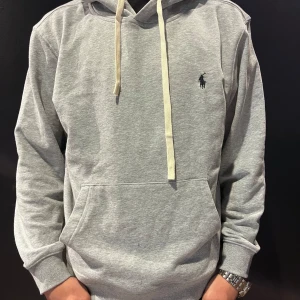 Grå hoodie från Polo Ralph Lauren - Säljer en klassisk grå hoodie från Polo Ralph Lauren med svart broderad logga på bröstet. Tröjan har huva med vita snören och en stor magficka. Perfekt för en avslappnad stil och enkel att matcha med jeans eller joggers. Tröjan är storlek M men skulle beskriva det mer som S.