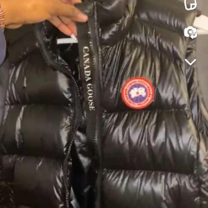 Svart dunväst från Canada Goose - Säljer en svart, glansig dunväst från Canada Goose med klassisk logotyp på bröstet och dragkedja framtill. Perfekt att bära över en tröja för extra värme. Snygg och tidlös design. Inga skador allt funkar som den ska, knappt använd prislappen finns ej kvar den är köpt från NK den är äkta finns andra bilder där prispallen syns.
