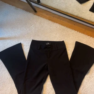 Svarta bootcut kostymbyxor med knappar - Säljer ett par svarta lågmidjade kostymbyxor i bootcut-modell med två knappar framtill och fickor med lock bak. Byxorna har en klassisk look och är perfekta för en stilren outfit. Aldrig använt dessa byxor då dem är för små. 