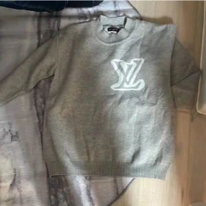Grå Louis Vuitton sweater - Säljer en grå stickad tröja från Louis Vuitton med stort vitt LV-logo på bröstet. Tröjan har rund halsringning och långa ärmar med ribbade muddar vid ärmslut och nederkant. Perfekt för en stilren och trendig look. Är i small men sitter som Large