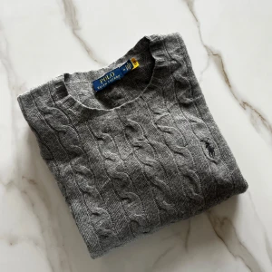 Ny grå kabelstickad tröja från Polo Ralph Lauren - Säljer en helt ny grå kabelstickad tröja från Polo Ralph Lauren. Tröjan har rund halsringning, långa ärmar och den ikoniska broderade loggan på bröstet. Perfekt för sommarkvällar och enkel att matcha med det mesta. Den är väldigt tunn alltså är den perfekt till sommaren! 90% Ull, 10% Kashmir.