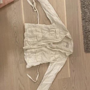 Vit linneskjorta med knytband från Neo Noir - Säljer en vit skjorta från Neo Noir i linne- och bomullsblandning. Skjortan har långa ärmar, två bröstfickor och knytband för knyta på ryggen och för en snygg passform.