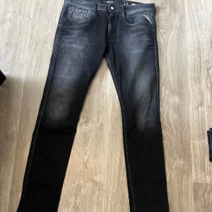 Svarta Replay Anbass jeans - Säljer ett par svarta Replay Anbass jeans med klassisk femficksdesign och snygga detaljer. Jeansen har normal passform och är tillverkade i stretchigt denim för extra komfort. Perfekta för dig som gillar stilrena och tidlösa jeans.