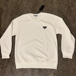 Vit sweatshirt från Prada - Säljer en stilren vit sweatshirt från Prada med diskret logga på bröstet. Tröjan är tillverkad i 100% bomull och har rund halsringning samt ribbade muddar vid ärmslut och nederkant. Perfekt för en clean och lyxig look.