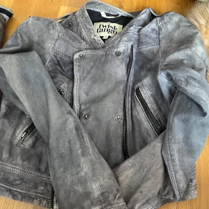 Grå skinnjacka från Twist & Tango - Säljer en grå skinnjacka från Twist & Tango med asymmetrisk dragkedja och flera snedställda fickor med dragkedjor. Jackan har långa ärmar och en cool, lite sliten look. Perfekt för dig som vill ha en edgy stil.