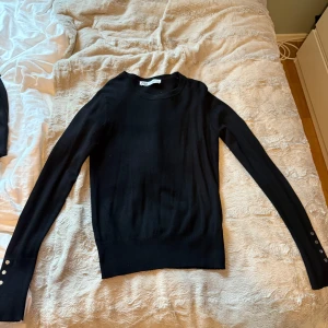 Svart långärmad tröja från Zara med knappar - Säljer en svart långärmad tröja från Zara i mjukt material. Tröjan har rund halsringning och dekorativa silvriga knappar vid ärmsluten. Enkel och stilren modell som passar till mycket.
