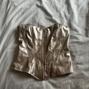 Guldig korsettopp från Amisu - Säljer en snygg guldig korsettopp i storlek XS. Toppen har dragkedja framtill och markerade sömmar som ger en cool och trendig look. Perfekt för att lyfta vilken outfit som helst!