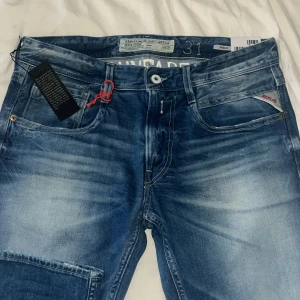 Blå Replay Anbass jeans - Säljer ett par snygga blå Replay Anbass jeans med slitningar och klassisk femficksdesign. Byxorna har en röd detalj vid fickan och tydlig Replay-logga. Modell ambass, helt nya(lapp kvar). Pris diskuterbart