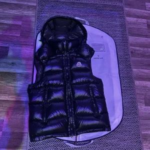 Svart Bormes dunväst - Säljer en svart glansig dunväst  med avtagbar huva och dragkedja framtill. Västen har två stora fickor och insidan har en etikett med tvättråd i serietidningsstil. Pris går att diskuteras och bara att höra av dig för flera frågor.