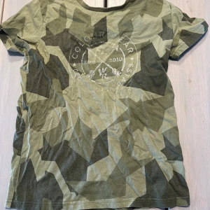 Grön camo t-shirt från Colour Wear - Säljer en grön camo t-shirt från Colour Wear med tryck på bröstet. T-shirten har korta ärmar och en klassisk rund halsringning. Perfekt för dig som gillar streetwear och vill sticka ut lite extra.