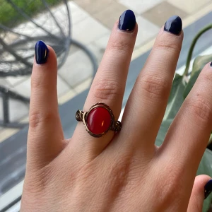 Ring med röd sten och bronsfärgad tråd - Unik ring med en stor oval röd sten infattad i antikguldfärgad metalltråd. Stenen är slät och blank, och tråden är konstnärligt virad runt stenen för en bohemisk känsla. Perfekt statement-piece för dig som gillar färg och vintageinspirerad stil.