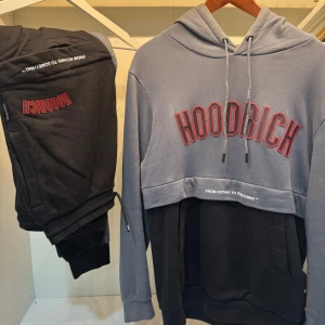 Grå och svart hoodie från Hoodrich - Säljer en snygg hoodie från Hoodrich i tvåfärgad design, grå upptill och svart nedtill. Stor logga i rött framtill och tryck på huvan. Tröjan har långa ärmar, huva med dragsko och mjukt material. Perfekt för en avslappnad streetwear-look.