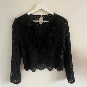 Svart spetsblus med volang från Zara - Säljer en svart spetsblus från Zara med långa ärmar och vackra volangdetaljer framtill. Blusen har ett genombrutet spetsmönster och knäppning i nacken. Perfekt för dig som gillar romantiska detaljer och stilren design.