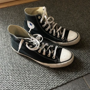 Svarta Converse All Star high-top sneakers - Klassiska svarta Converse All Star high-top sneakers med vit tå och vita snören. Skorna har den ikoniska loggan på sidan och en platt sula. Perfekta för en avslappnad och cool stil. Bra skick! Hör av dig vid funderingar!
