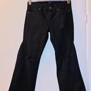 Svarta bootcut jeans med slitningar - Svarta jeans med bootcut passform och slitningar vid knäna. Klassisk femficksmodell med knapp och dragkedja framtill. Perfekta för en avslappnad och trendig look.