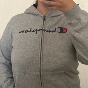Grå zip-hoodie från Champion - Säljer en grå hoodie med dragkedja från Champion. Klassisk modell med marinblå färg inuti huvan och fickor framtill. Stor logga på bröstet. XL i barnstorlek men skulle passa XS/S. 