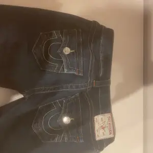 Säljer ett par mörkblå jeansbyxor från True Religion med klassiska kontrastsömmar och logga på bakfickan. Byxorna har låg midja och utsvängda ben, samt detaljerade fickor med knappar. Perfekta för dig som gillar en tidlös jeansstil.