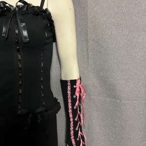 Svarta handskar med rosa snörning - Unika svarta handskar med dekorativ rosa snörning och spetsdetaljer längs kanterna. Perfekta för att ge en extra touch till din outfit. Snörningen ger en justerbar passform och en elegant look. Helt nya i bomullstrikå.
