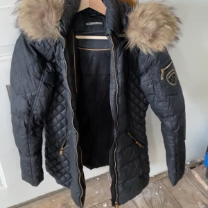 Svart quiltad dunjacka med fuskpäls från Rockandblue - Svart quiltad dunjacka från Rockandblue med stor huva kantad av beige fuskpäls. Jackan har guldiga dragkedjor, två fickor fram och Rockandblue-logga på ärmen. Snygg och varm modell med lång ärm och figurnära passform. Finns ett litet hål på armen. Syns på sista bilden. Kostar 4000 typ ny 