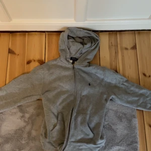 Grå hoodie från Polo Ralph Lauren - Grå hoodie från Polo Ralph Lauren med dragkedja framtill och huva. Klassisk design med liten broderad logga på bröstet och två fickor framtill. Perfekt för en avslappnad stil.