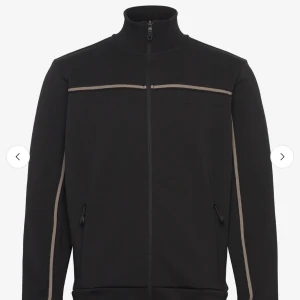 Svart trackjacka med beige detaljer från BOSS - Säljer en svart trackjacka från BOSS med diskreta beige linjer över bröstet och längs ärmarna. Jackan har hög krage, hel dragkedja och två fickor med dragkedja framtill. Perfekt för en sportig och stilren look.