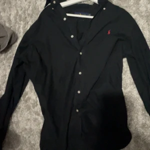 Svart skjorta från Ralph Lauren - Snygg svart skjorta från Ralph Lauren i klassisk passform. Skjortan har knappar framtill och en liten röd broderad logga på bröstet. Perfekt för en stilren look.