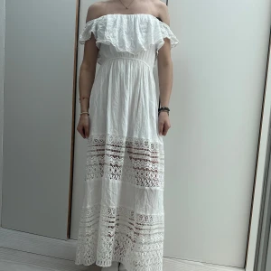 Vit offshoulder klänning med spetsdetaljer - Säljer en romantisk vit offshoulder klänning med vackra spetsdetaljer och volang upptill. Klänningen är lång och perfekt för sommarens festligheter. Den har en luftig passform och är tillverkad i ett lätt material som ger en skön känsla. 