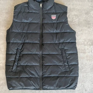 Svart dunväst från Gant - Snygg svart dunväst från Gant med dragkedja och ett stilrent märke på bröstet. Perfekt för kyligare dagar och ger en sportig look. Västen har en quiltad design och praktiska fickor.