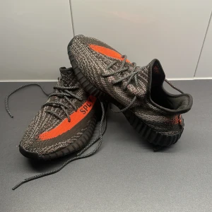 Adidas Yeezy Boost 350 V2 - Snygga Adidas Yeezy Boost 350 V2 med en unik design i grått och orange. Skorna har en stickad ovandel med snörning och en bekväm Boost-sula. Perfekta för dig som vill ha en stilren och modern look.
