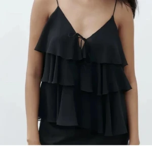 Svart volangtopp från Zara - Snygg svart topp från Zara med tunna axelband och flera lager av volanger. Toppen har en knytning framtill som ger en extra detalj. Perfekt för en stilren look. Använd en gång🩷