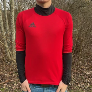 Röd långärmad tröja från Adidas - Säljer en snygg röd långärmad tröja från Adidas med svart logga på bröstet. Perfekt för sportiga aktiviteter eller en casual look. Tröjan har en bekväm passform och är tillverkad i ett lätt material.