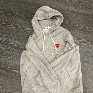 Grå hoodie från Comme des Garçons - Säljer en stilren grå hoodie från Comme des Garçons med det ikoniska röda hjärtat broderat på bröstet. Tröjan har en justerbar huva med vita snören och en bekväm passform. Perfekt för en avslappnad look.