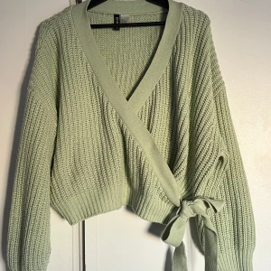 Grön omlottkofta från H&M - Säljer en mysig grön omlottkofta från H&M Divided. Koftan är stickad med ett ribbat mönster och har långa ärmar. Den har en knytning i midjan för en snygg passform. 