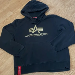 Svart hoodie från Alpha Industries - Säljer en svart hoodie från Alpha Industries med guldtryck på bröstet. Hoodien har en klassisk känguruficka och justerbar huva med dragsko. Tillverkad i en bomulls- och polyesterblandning för extra komfort. Perfekt för en avslappnad stil.