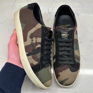 Saint Laurent camo skor - Snygga dojor från Saint Laurent i ett coolt camouflage-mönster. Skorna har svarta snören och en vit sula som ger en stilren kontrast. Perfekta för en avslappnad och trendig look.