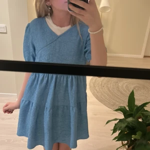 Blå klänning från Lindex - Säljer en söt blå klänning från Lindex med korta ärmar och omlott-design. Klänningen har en fin struktur och är perfekt för sommarens varma dagar. Den är gjord i ett mjukt material som känns skönt mot huden.