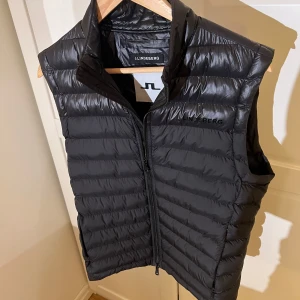 Svart väst från J.Lindeberg - Snygg svart, lite glänsig dunväst från J.Lindeberg med dragkedja framtill. Perfekt nu till våren. Västen har en stilren design med diskret logga på bröstet. Köpt för 1800 kr och använd endast ett fåtal gånger! Skriv gärna vid minsta fundering!🤗