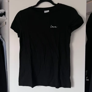 Svart t-shirt med broderad text - Säljer en stilren svart t-shirt från H&M Divided med en liten vit broderad text 'love' på bröstet. T-shirten har korta ärmar och en klassisk rund halsringning. Perfekt för en avslappnad look 🖤 Önskar du veta exkta mått? Tveka inte att höra av dig! 