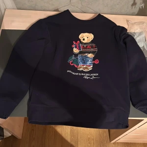 Mörkblå tröja med Polo Bear från Ralph Lauren - Säljer en mörkblå tröja från Ralph Lauren med det ikoniska Polo Bear-motivet. Björnen har en julig outfit med en present i handen. Perfekt för vintersäsongen och julfirandet. Tröjan har långa ärmar och en klassisk passform.scick 9/10 säljs på grund ut av att den har blivit för liten.Priset är ej hugget i sten