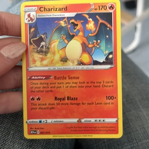 Charizard Pokémonkort - Charizard-kort med 170 HP och förmågan Battle Sense. Attacken Royal Blaze gör 100+ skada och ökar med 50 för varje Leon-kort i din hög. Kortet är nummer 001/015.