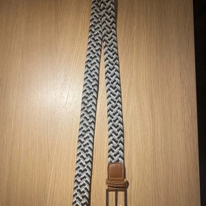Vävt bälte med läderdetaljer - Snyggt vävt bälte i svart och vitt med bruna läderdetaljer. Perfekt för att ge en stilren touch till din outfit. Hör av dig vid funderingar! 