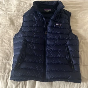 Blå dunväst från Patagonia - Säljer en snygg blå dunväst från Patagonia. Västen har en dragkedja framtill och två praktiska fickor med dragkedjor. Perfekt för kyligare dagar och ger en stilren look. Västen kostar 2299kr ny, mitt pris 850kr. Det inte ett spikat pris!!