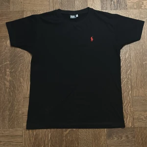 Svart t-shirt från Polo Ralph Lauren - Snygg svart t-shirt från Polo Ralph Lauren med det klassiska röda broderade logotypen på bröstet. Perfekt för en stilren look. T-shirten har korta ärmar och är tillverkad i mjukt material för extra komfort.